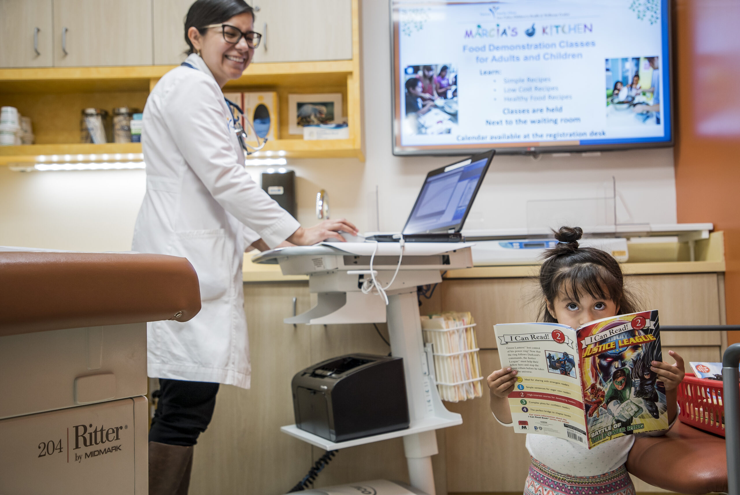 A female doctor smiles near a computer in a medical exam room while a young girl sits on an exam table, reading a comic book and looking at the camera. A screen in the background displays class information.