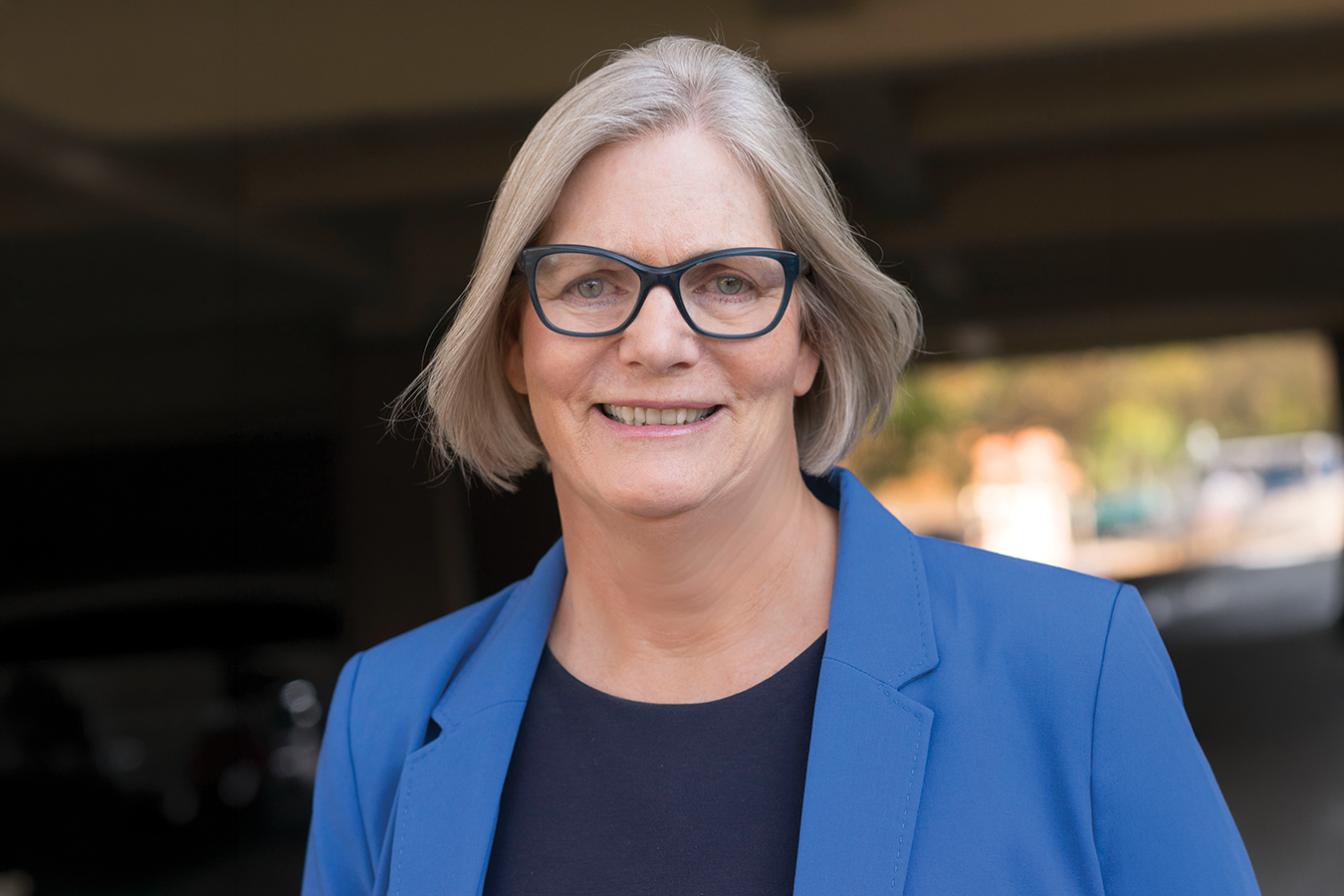 A Smiling Older Woman With Gray Hair And Glasses Wears A Blue Blazer Over A Dark Top, Standing Outdoors With A Blurred Background.