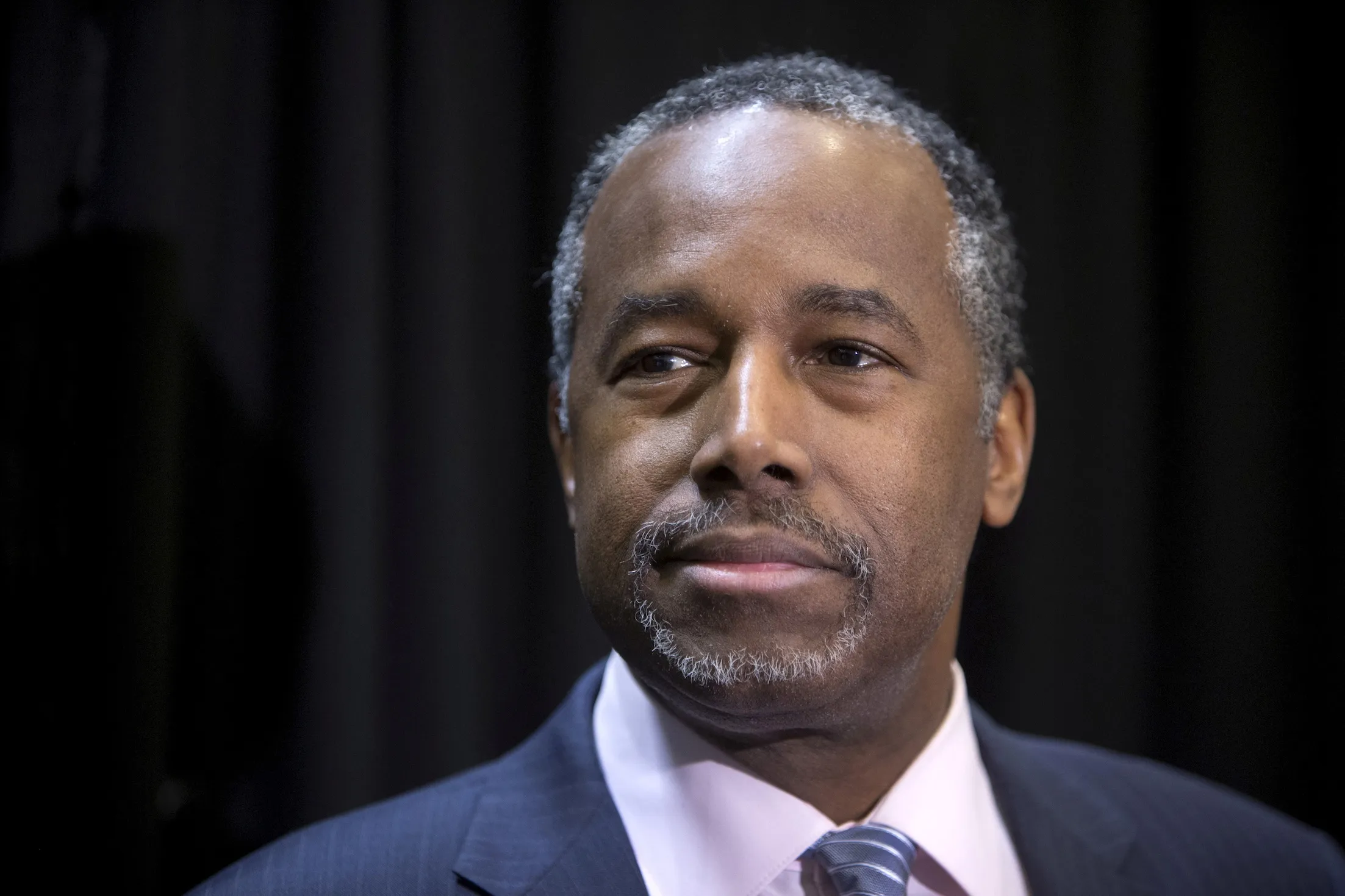 A Middle-aged Man With Short, Graying Hair And A Trimmed Mustache And Beard Is Wearing A Dark Suit, Light Pink Shirt, And Striped Tie, Standing In Front Of A Dark Background, Looking Slightly To The Side. A Middle-aged Man With Short, Graying Hair And A Trimmed Mustache And Beard Is Wearing A Dark Suit, Light Pink Shirt, And Striped Tie, Standing In Front Of A Dark Background, Looking Slightly To The Side.