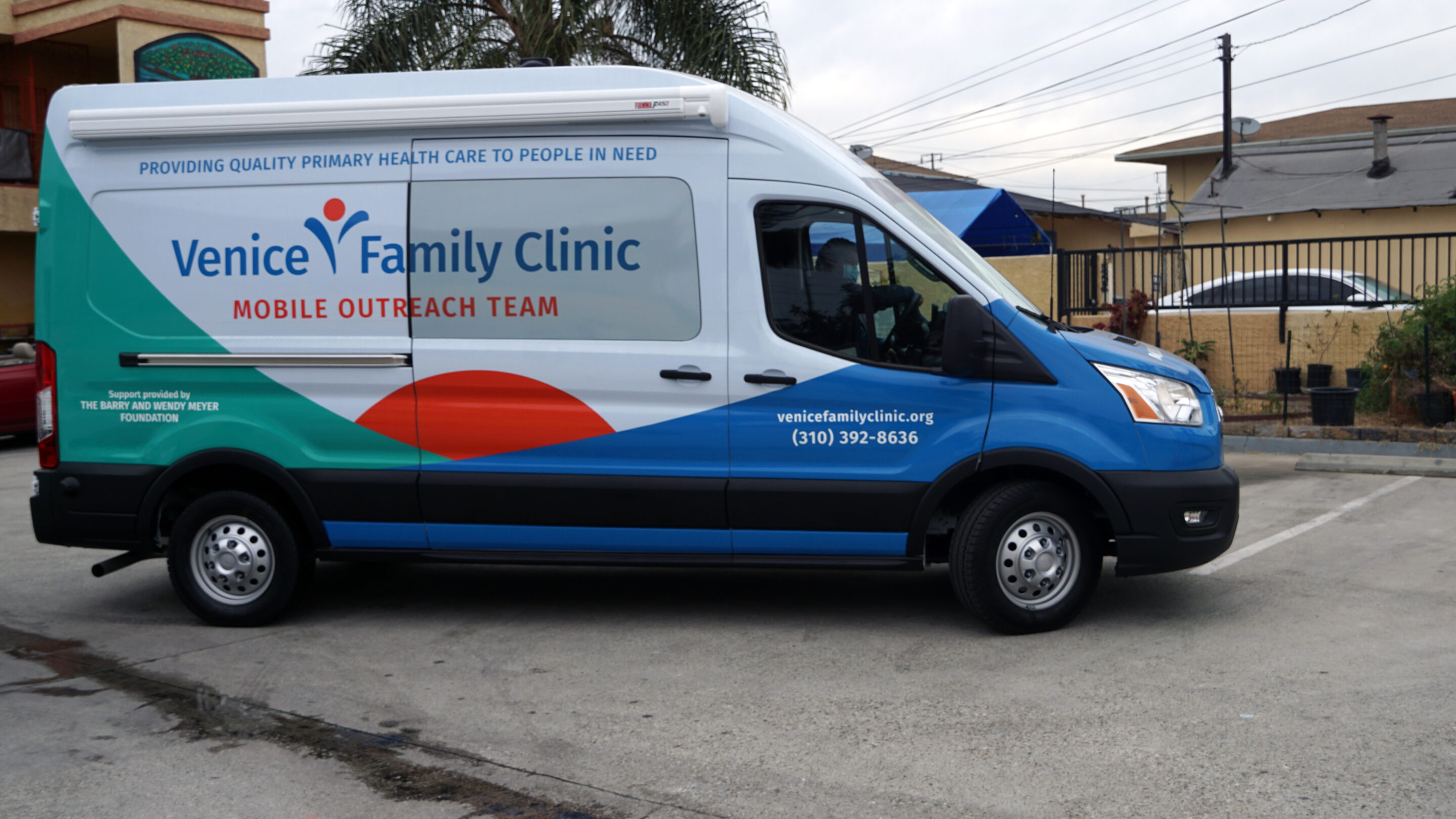 A Venice Family Clinic Mobile Outreach Van Is Parked On A Street. The Van Is White And Blue With Clinic Branding And Contact Information, And Is Parked Near A Fenced Building And Palm Tree. A Venice Family Clinic Mobile Outreach Van Is Parked On A Street. The Van Is White And Blue With Clinic Branding And Contact Information, And Is Parked Near A Fenced Building And Palm Tree.