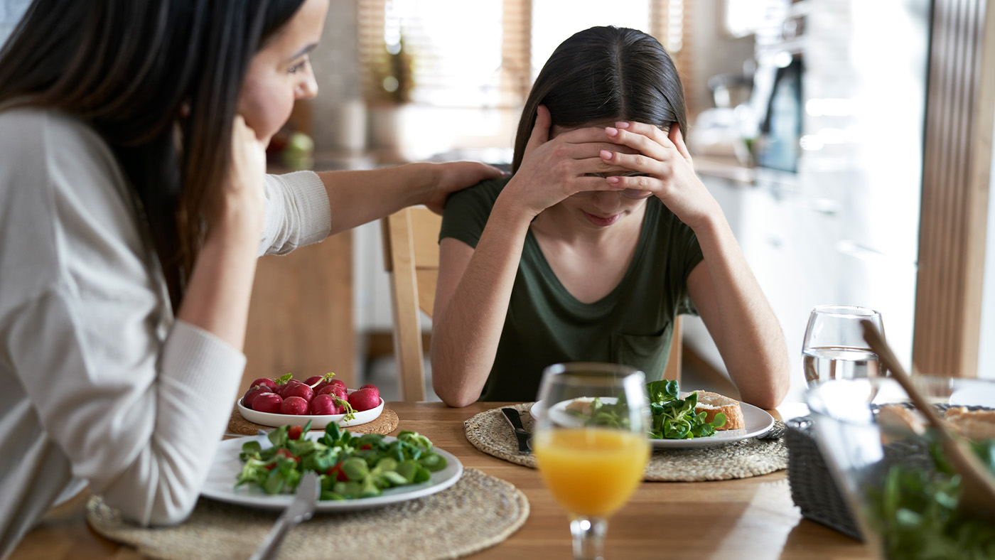 Two People Sit At A Table With Salad And Juice. One Person Comforts The Other, Who Has Her Head In Her Hands And Appears Upset. The Scene Is Set In A Bright Kitchen.