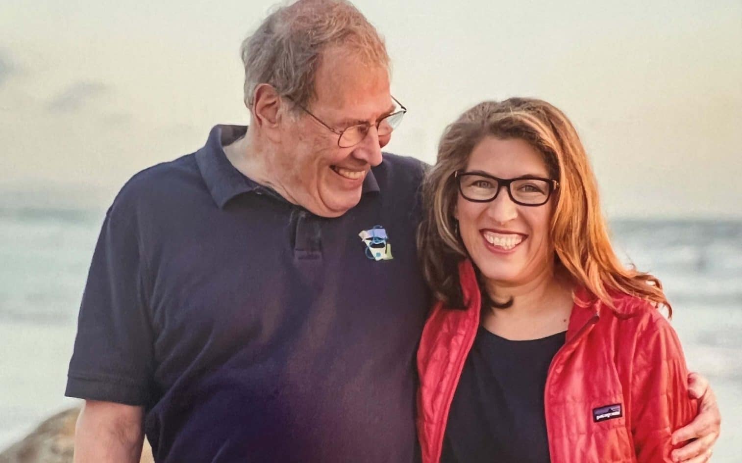 An Older Man In A Navy Polo Shirt And A Woman In A Red Jacket Stand Close Together, Smiling And Looking Happy, With The Beach And Ocean In The Background.