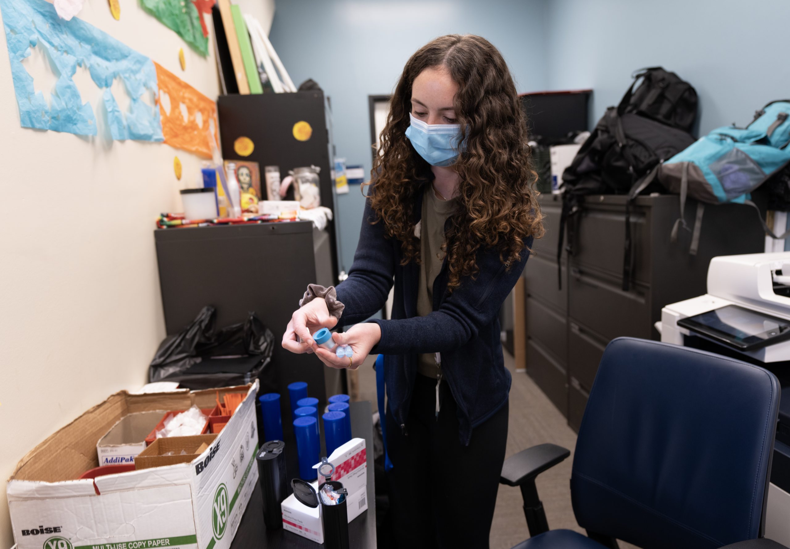 A Woman Wearing A Face Mask Stands In An Office, Holding And Arranging Items Near A Box On A Table. Behind Her Are Shelves, Stacks Of Papers, And Various Office Supplies. A Woman Wearing A Face Mask Stands In An Office, Holding And Arranging Items Near A Box On A Table. Behind Her Are Shelves, Stacks Of Papers, And Various Office Supplies.