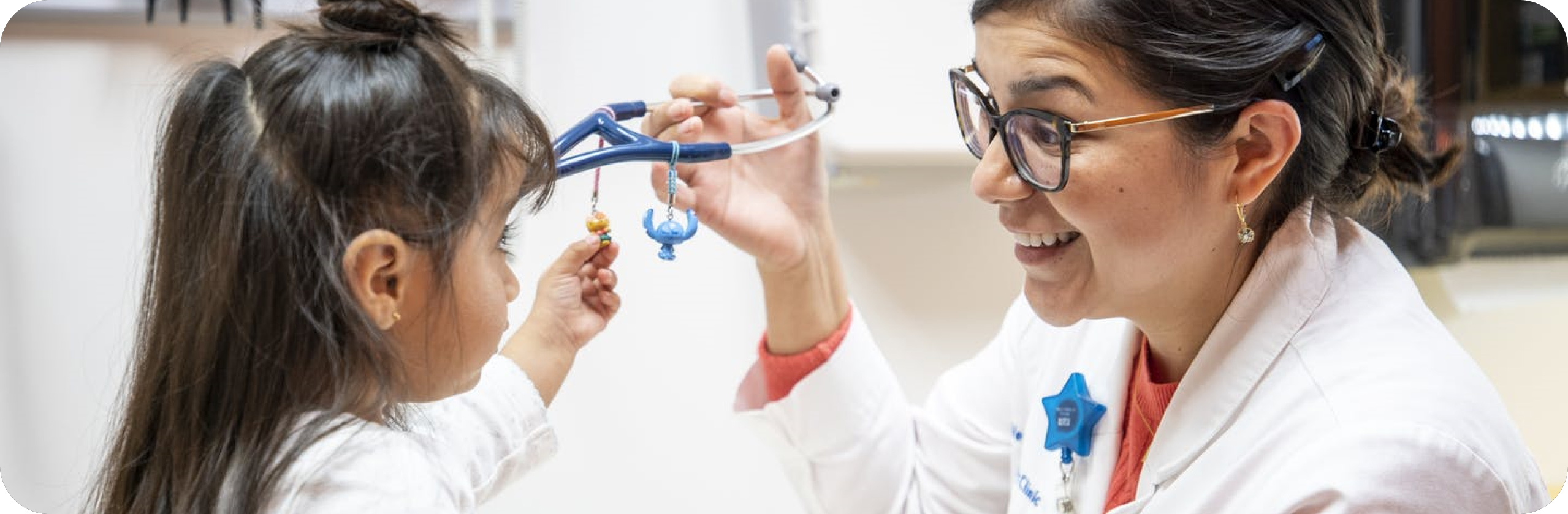 A female doctor smiling and holding a stethoscope while interacting with a young girl who is reaching for the stethoscope toys, in a brightly-lit medical office.