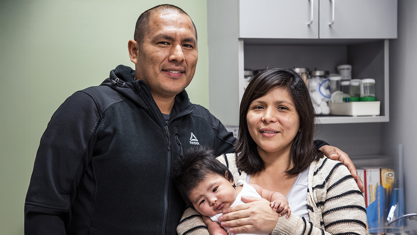 Una pareja sonriente sostiene en brazos a su bebé en una sala de reconocimiento médico, de pie frente a armarios con material médico. Una pareja sonriente sostiene en brazos a su bebé en una sala de reconocimiento médico, de pie frente a armarios con material médico.