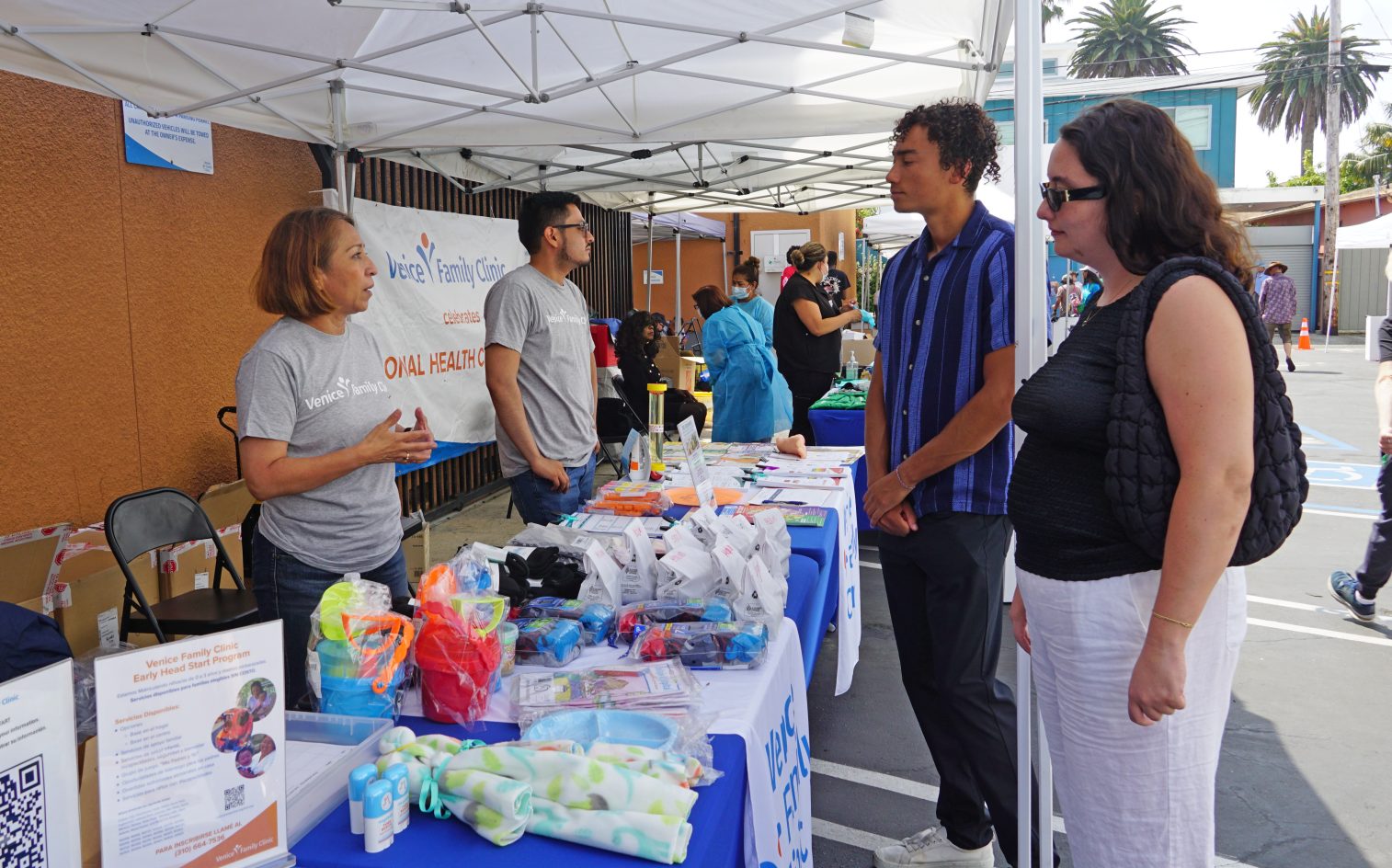 Un grupo de personas conversa en un puesto de una feria de salud al aire libre con folletos informativos, kits de higiene y juguetes bajo un toldo blanco. Al fondo se ven otros puestos y visitantes. Un grupo de personas conversa en un puesto de una feria de salud al aire libre con folletos informativos, kits de higiene y juguetes bajo un toldo blanco. Al fondo se ven otros puestos y visitantes.