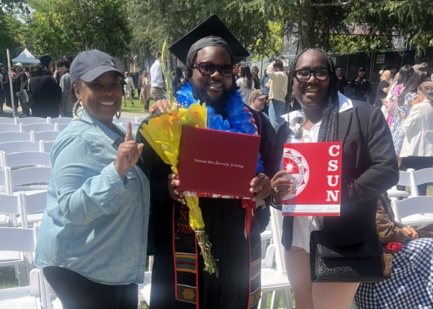 Tres personas sonríen en una graduación al aire libre. El graduado, con toga y birrete, sostiene un diploma y un ramo de flores. Las dos mujeres a su lado sostienen un cartel de CSUN y hacen gestos con un número uno. Al fondo, sillas blancas y personas. Tres personas sonríen en una graduación al aire libre. El graduado, con toga y birrete, sostiene un diploma y un ramo de flores. Las dos mujeres a su lado sostienen un cartel de CSUN y hacen gestos con un número uno. Al fondo, sillas blancas y personas.