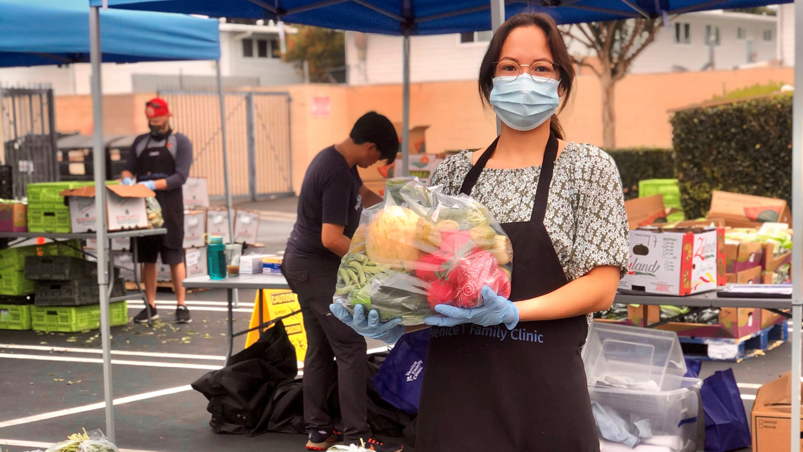 Una mujer con mascarilla, guantes y delantal sostiene una bolsa de productos frescos en un acto de distribución de alimentos al aire libre. Al fondo, otros voluntarios clasifican cajas de alimentos bajo carpas azules.