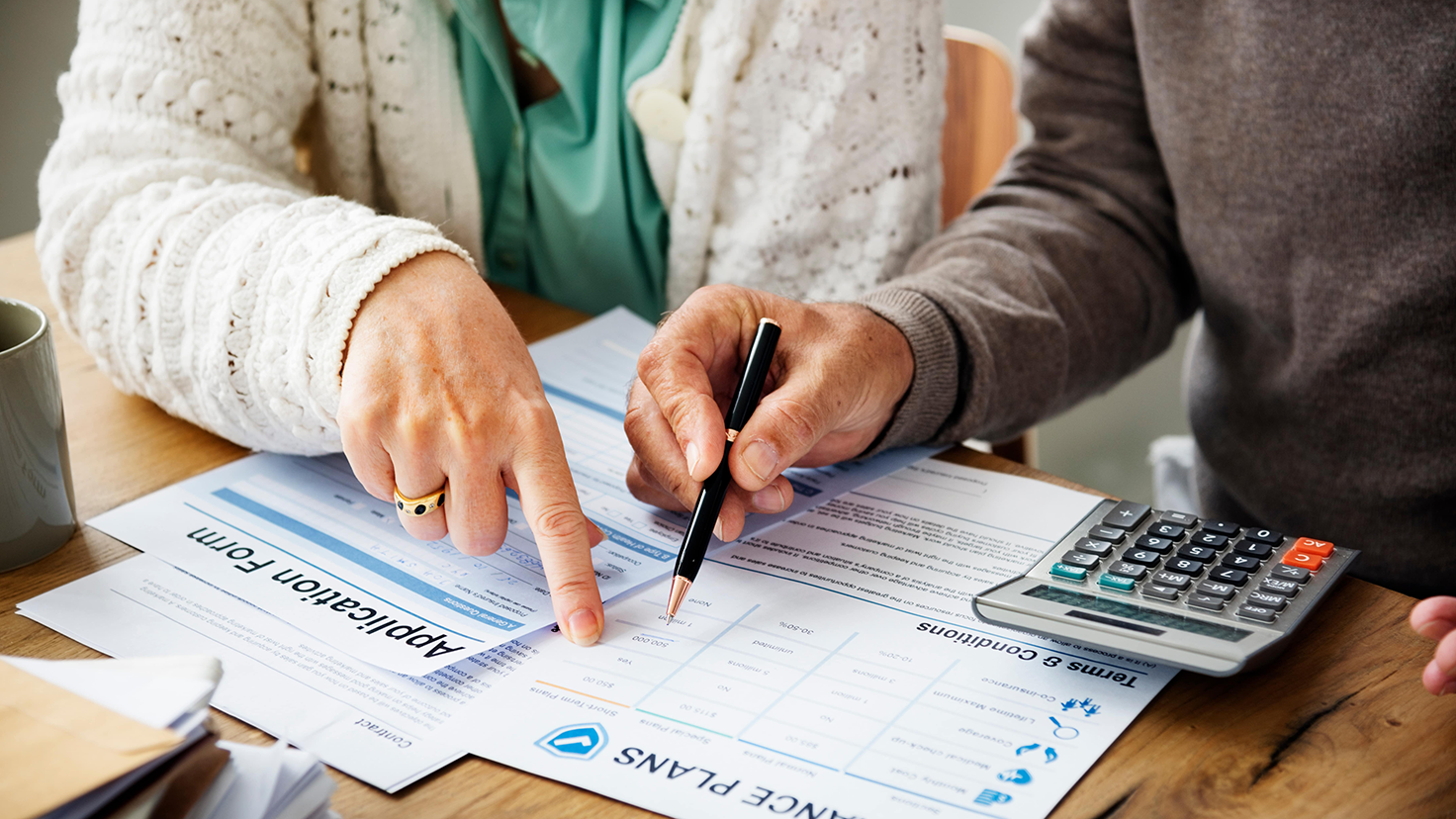 Dos personas revisan el papeleo del seguro en una mesa. Una señala una sección de un formulario de solicitud mientras la otra sostiene un bolígrafo y una calculadora, discutiendo detalles o cálculos relacionados con los formularios.