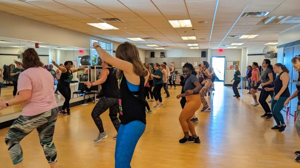 Un grupo de personas participa en una clase de fitness dance en un luminoso estudio con espejos a lo largo de una pared y suelos de madera. Todo el mundo está en pleno movimiento, siguiendo al instructor que va delante.