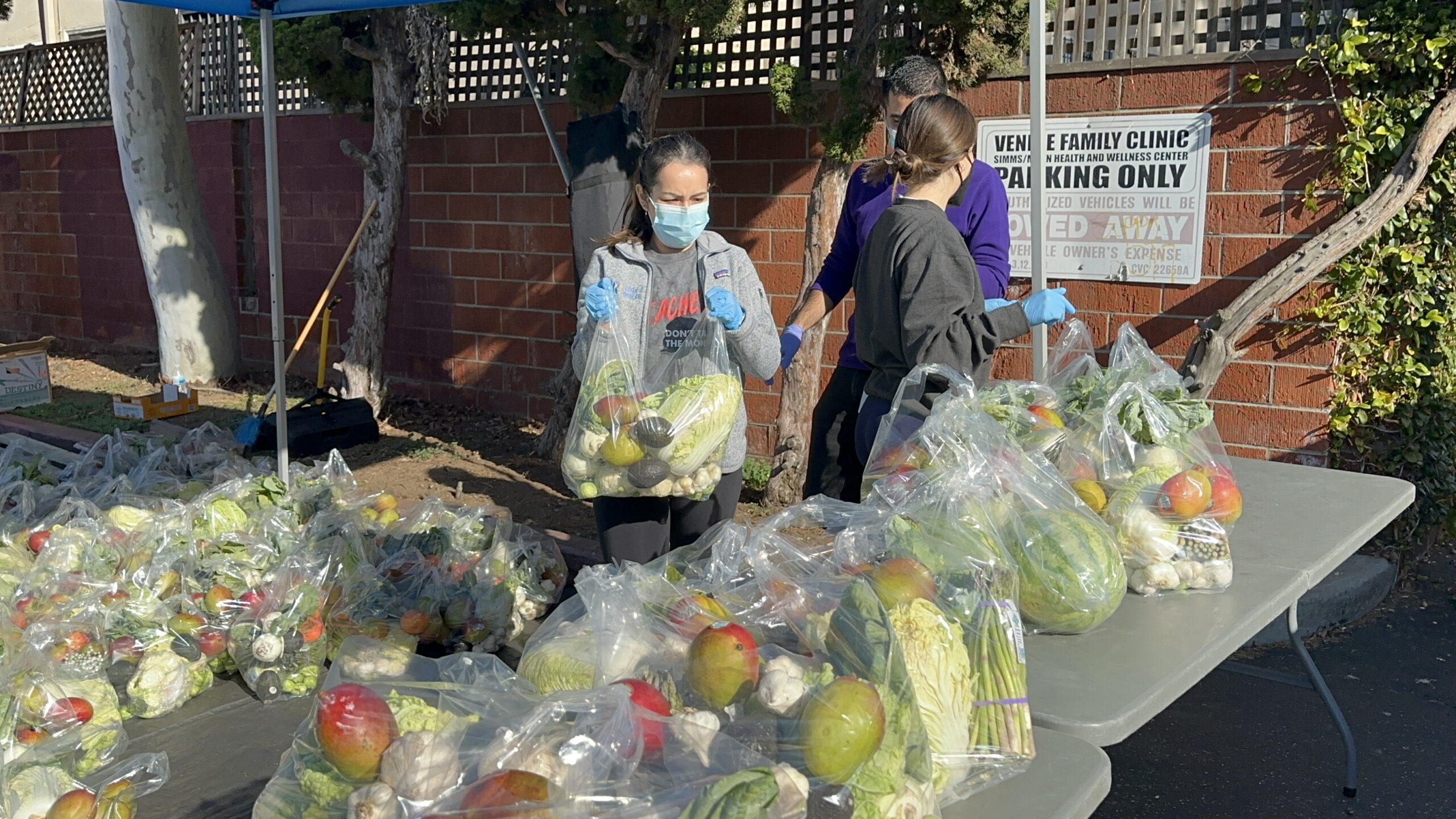 Personas con mascarillas y guantes distribuyen bolsas de productos frescos en una mesa de donación de alimentos al aire libre, con varias bolsas llenas de frutas y verduras colocadas en mesas bajo un toldo.