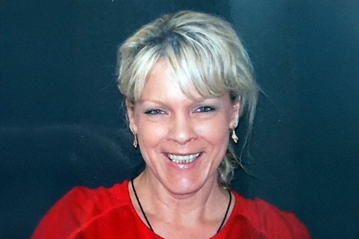 A smiling woman with blonde hair wearing a red top and small earrings, posed against a plain dark background.