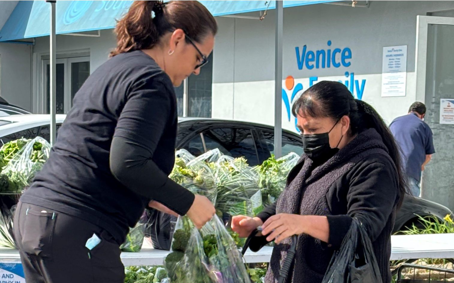A Woman Hands A Bag Of Leafy Greens To Another Woman Wearing A Mask And Holding A Reusable Shopping Bag At An Outdoor Market Stand. A Building With A “Venice Family” Sign Is Visible In The Background.