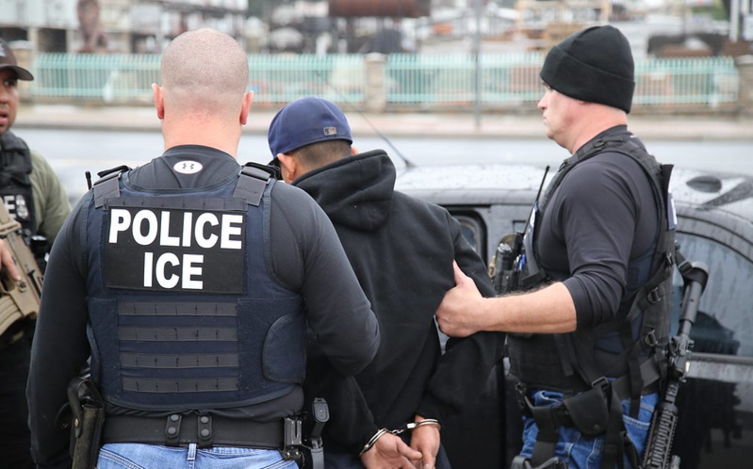 Several Law Enforcement Officers Wearing “POLICE ICE” Vests Detain A Handcuffed Person In A Black Hoodie Next To A Black Vehicle On A City Street.