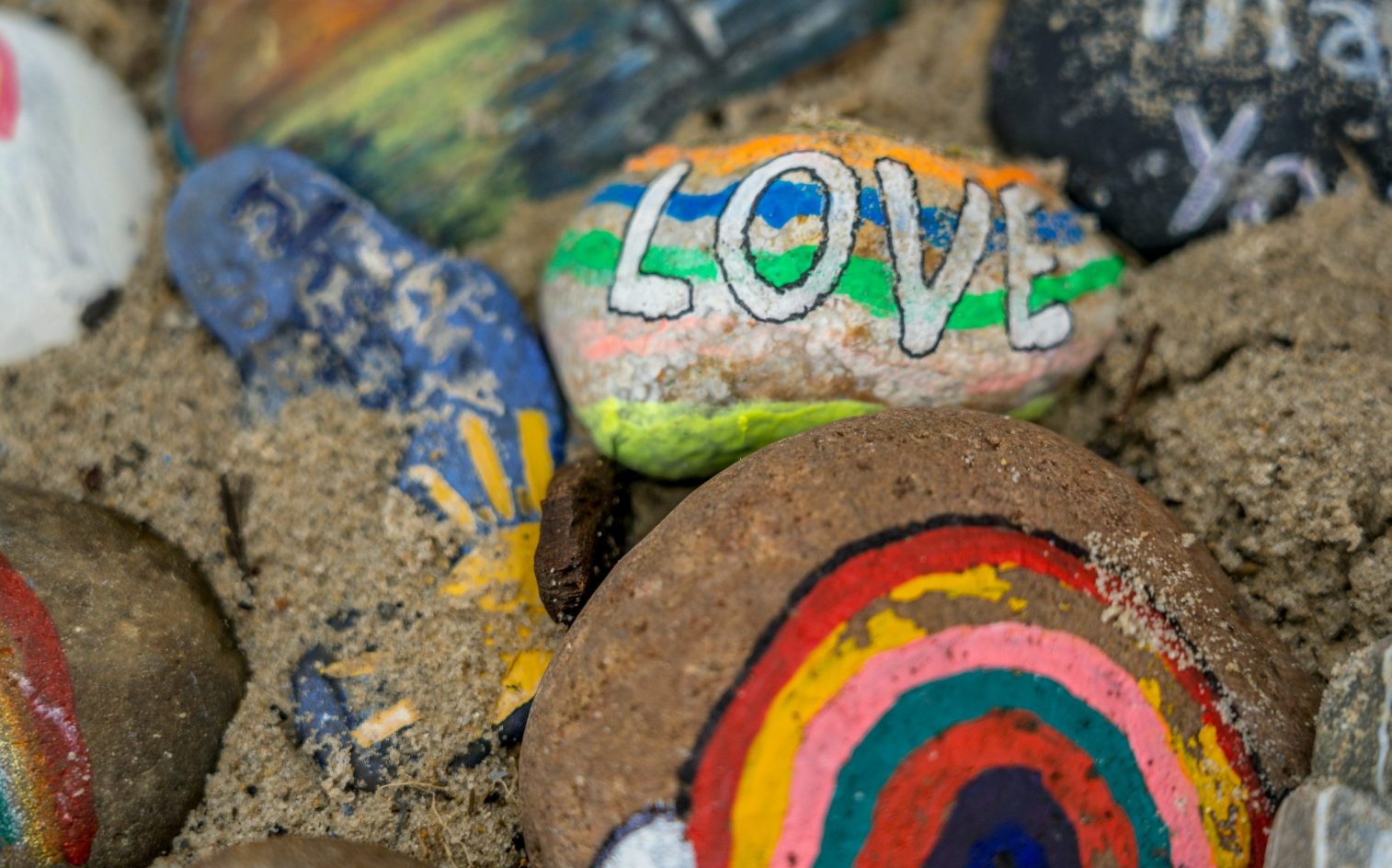 Coloridas rocas pintadas sobre la arena, incluida una con la palabra LOVE en letras gruesas con rayas arco iris y otra con un diseño arco iris. Coloridas rocas pintadas sobre la arena, incluida una con la palabra LOVE en letras gruesas con rayas arco iris y otra con un diseño arco iris.