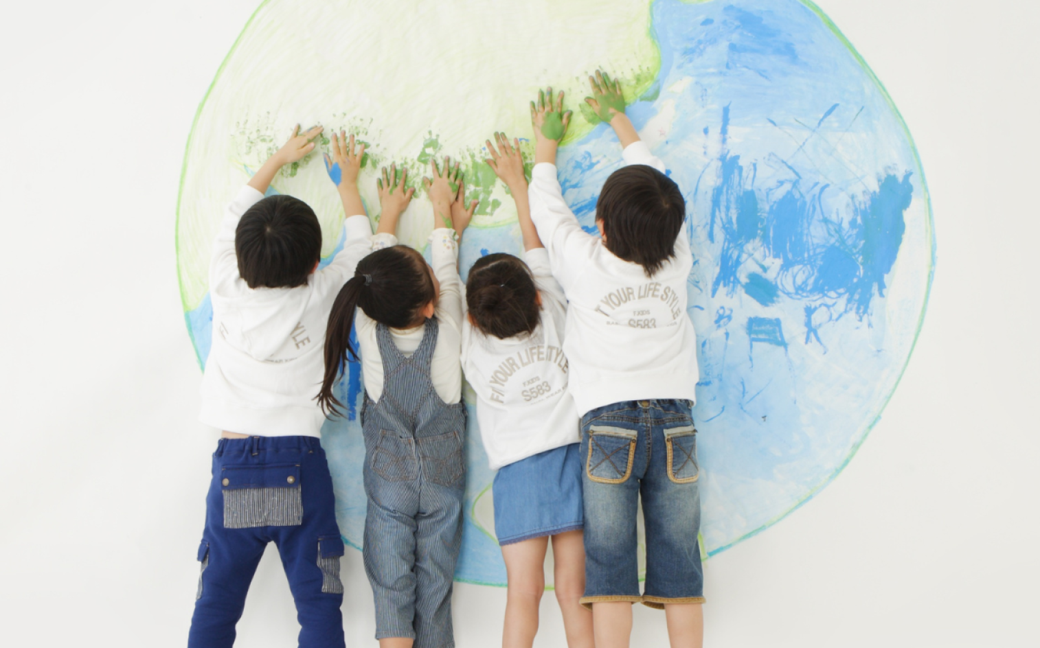 Four Children With Their Backs To The Camera Press Their Green-painted Hands Onto A Large Drawing Of Earth On A Wall, Wearing White Tops And Blue Denim Bottoms. Four Children With Their Backs To The Camera Press Their Green-painted Hands Onto A Large Drawing Of Earth On A Wall, Wearing White Tops And Blue Denim Bottoms.