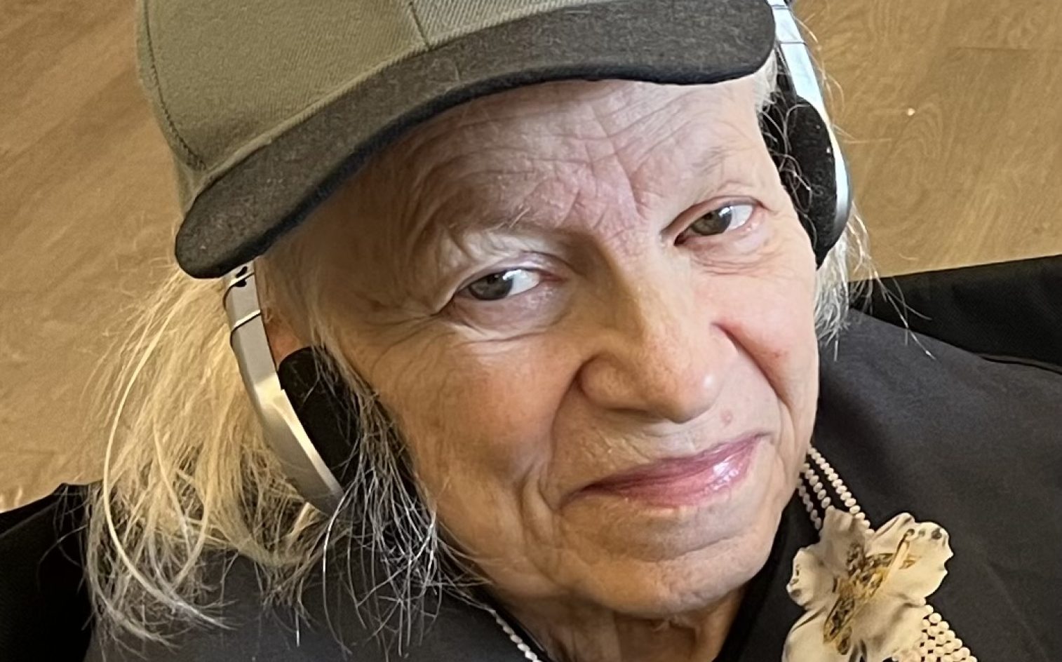 An older woman with long white hair wears a gray baseball cap and over-ear headphones. She smiles gently at the camera and is dressed in a dark top with a flower decoration.