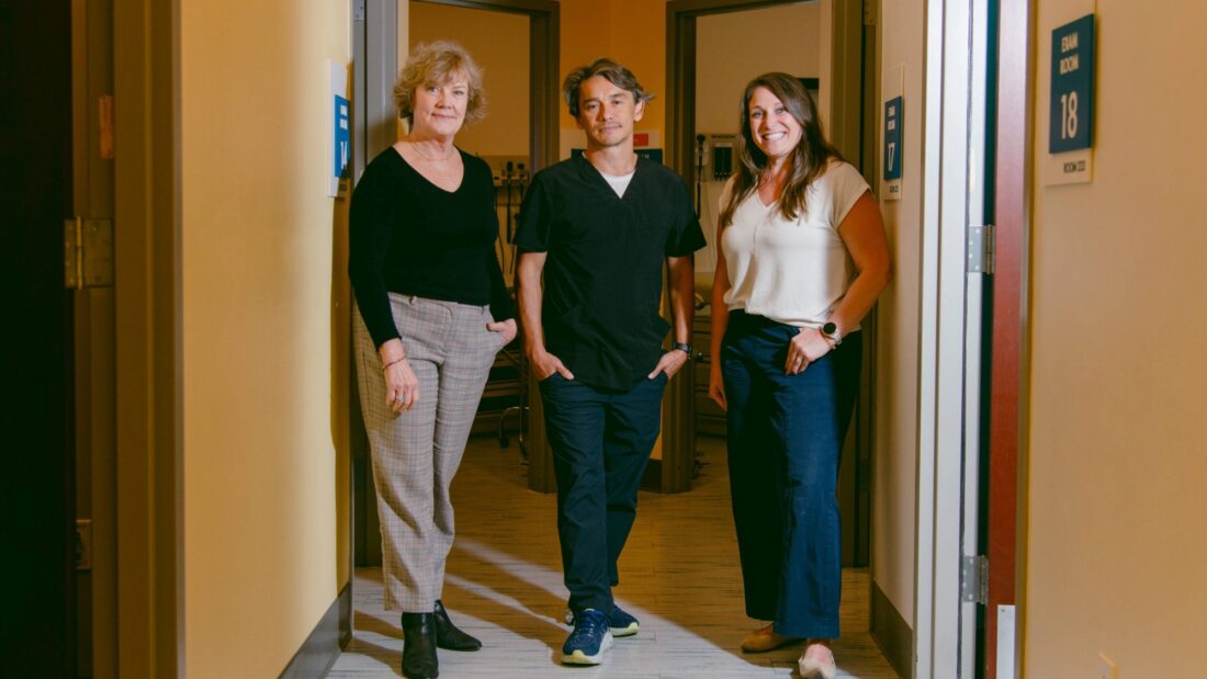 Three People Stand In A Hallway Between Two Open Doors Labeled 18 And 17. They Are Smiling And Dressed In Business-casual Attire, With Warm Yellow Lighting Illuminating The Scene.