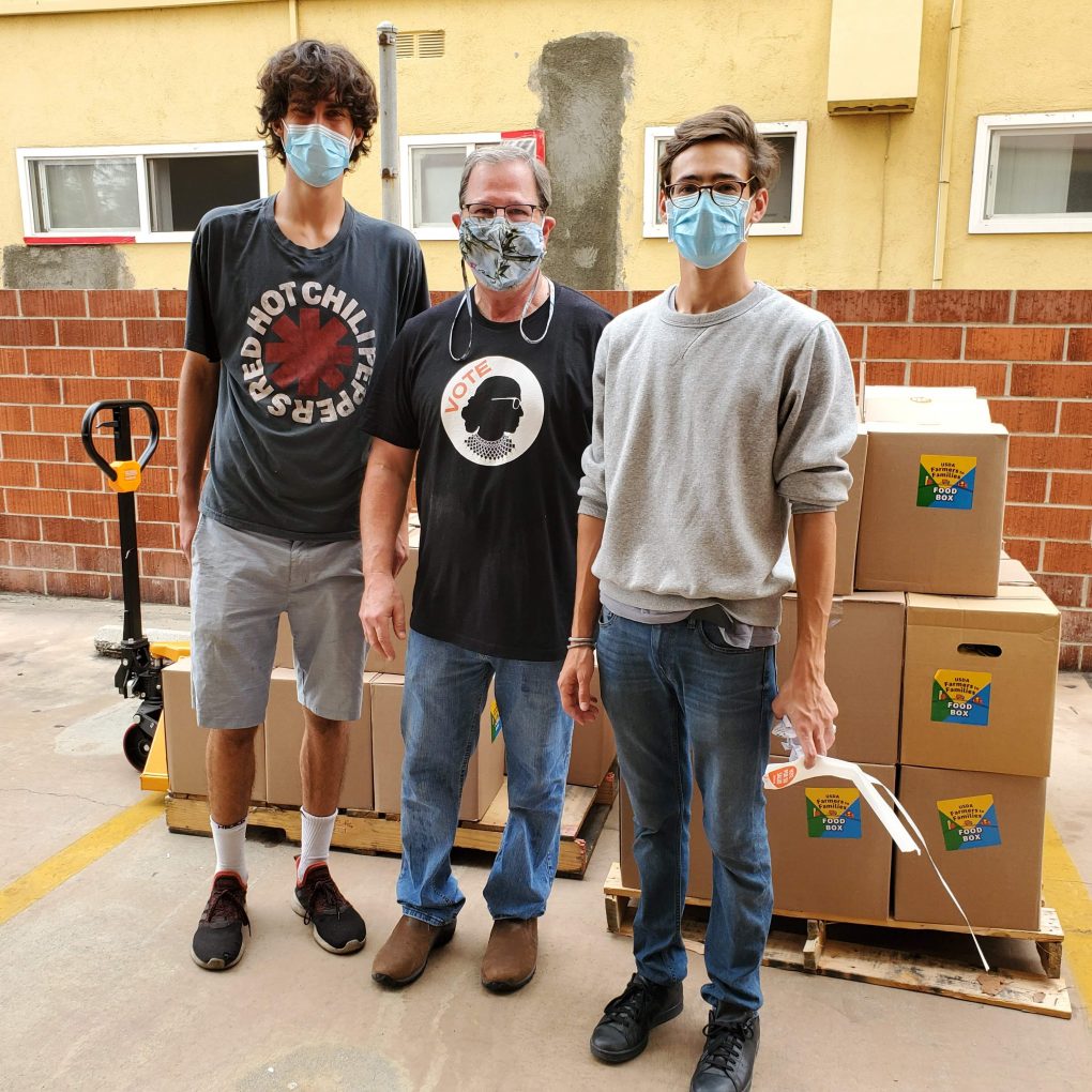 Three People Wearing Face Masks Stand In Front Of Stacked Boxes On Pallets Outside A Brick Building. They Appear To Be Volunteers Or Workers Involved In A Distribution Or Relief Effort.