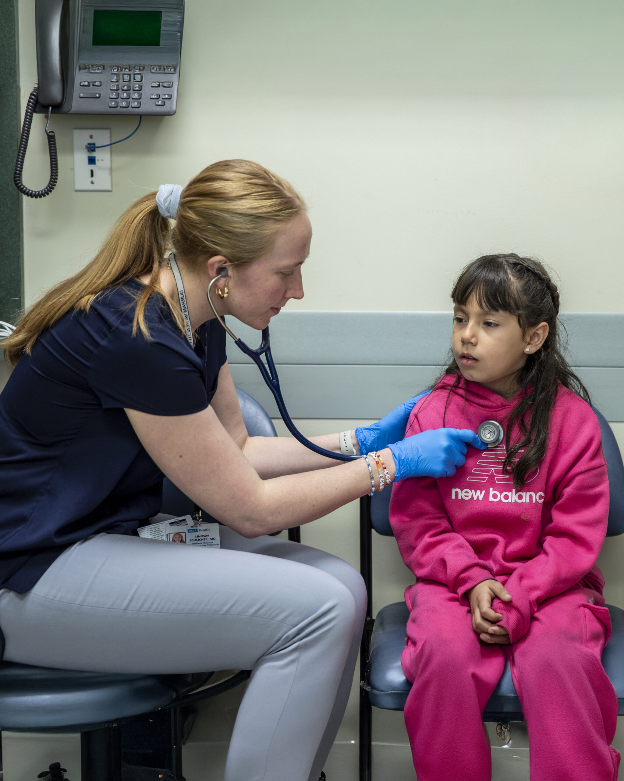 A Healthcare Worker Uses A Stethoscope To Examine A Young Girl In A Pink Tracksuit During A Medical Checkup. They Are Seated In A Clinic Room, And The Healthcare Worker Wears Blue Gloves.