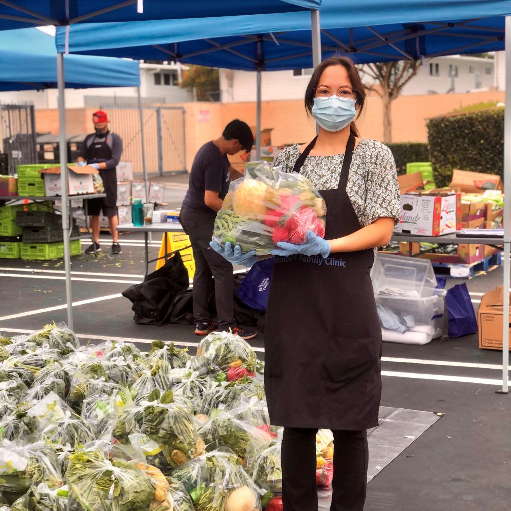 A woman wearing a mask, gloves, and apron stands under a blue canopy, holding a bag of fresh produce. Behind her, others sort vegetables and food supplies into bags at an outdoor distribution event.