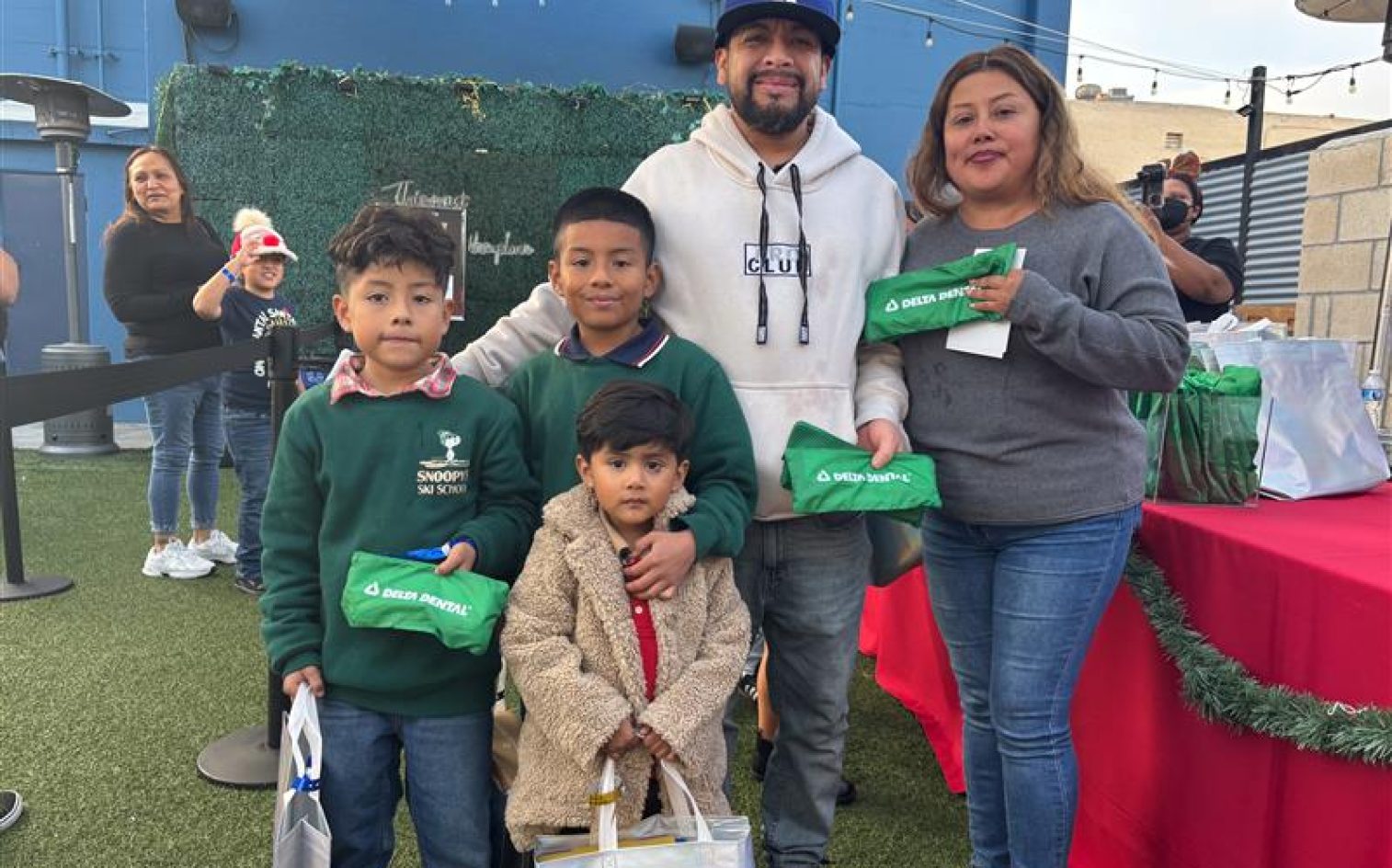 A Family Of Five Poses Outdoors At An Event. The Parents Stand Behind Three Children Who Are Holding Goodie Bags. Festive Decorations And A Table With Wrapped Gifts Appear In The Background. A Family Of Five Poses Outdoors At An Event. The Parents Stand Behind Three Children Who Are Holding Goodie Bags. Festive Decorations And A Table With Wrapped Gifts Appear In The Background.