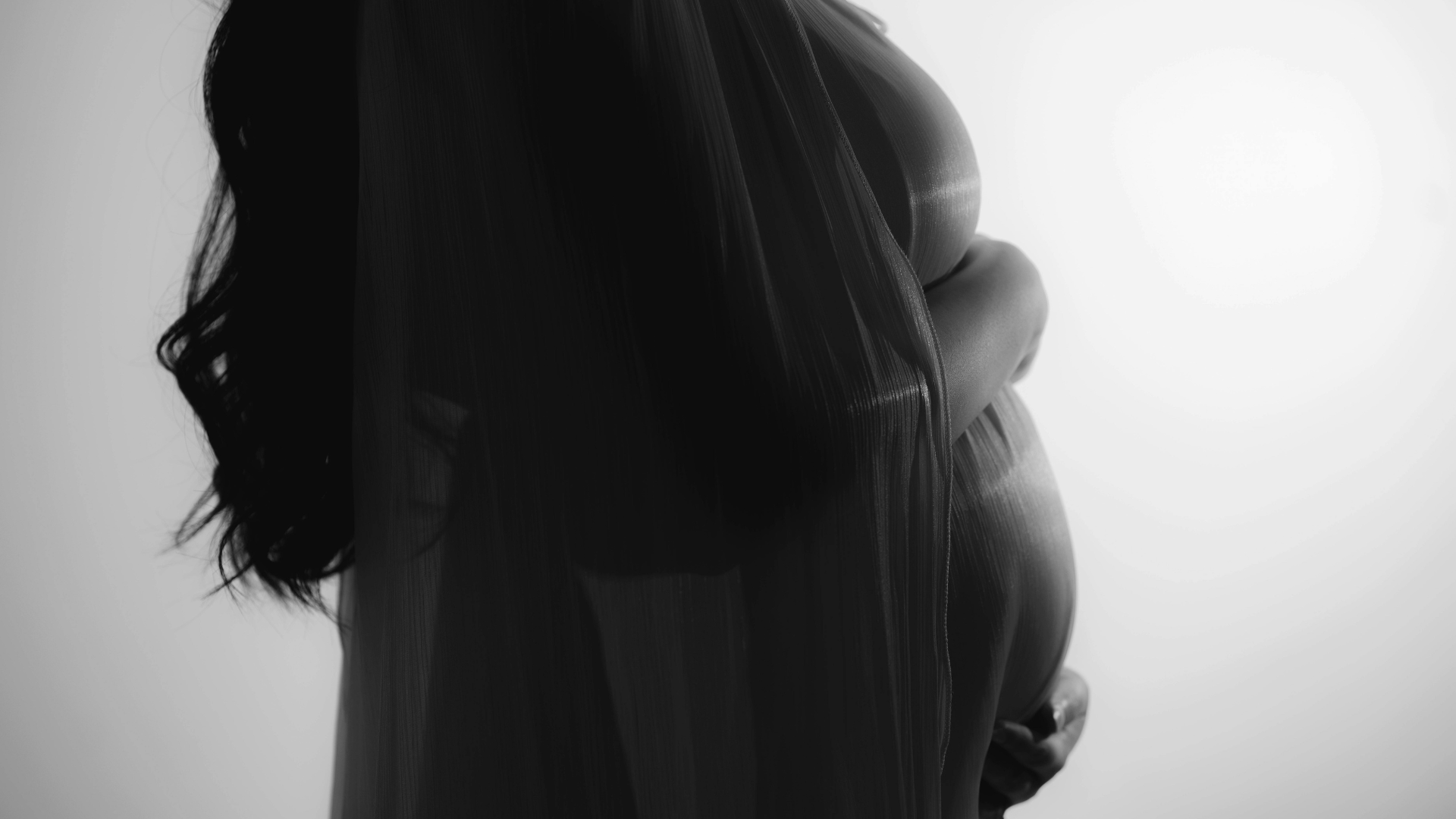 A Black And White Photo Of A Pregnant Woman In Profile, Draped In Sheer Fabric, Gently Holding Her Belly Against A Light Background. A Black And White Photo Of A Pregnant Woman In Profile, Draped In Sheer Fabric, Gently Holding Her Belly Against A Light Background.