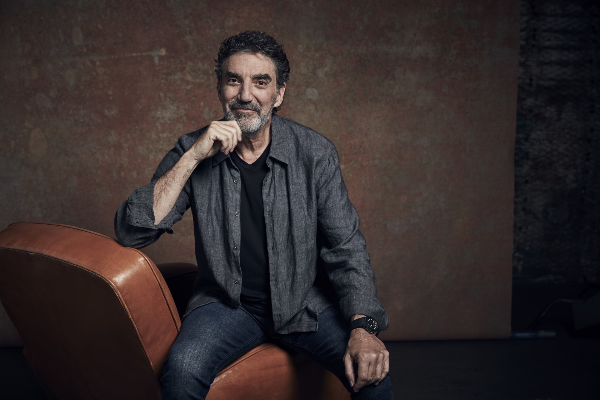 A Man With Gray Hair And A Beard Sits On A Brown Leather Chair, Wearing A Gray Shirt Over A Black T-shirt And Jeans. He Smiles Slightly And Rests His Chin On His Hand Against A Plain Brown Background.