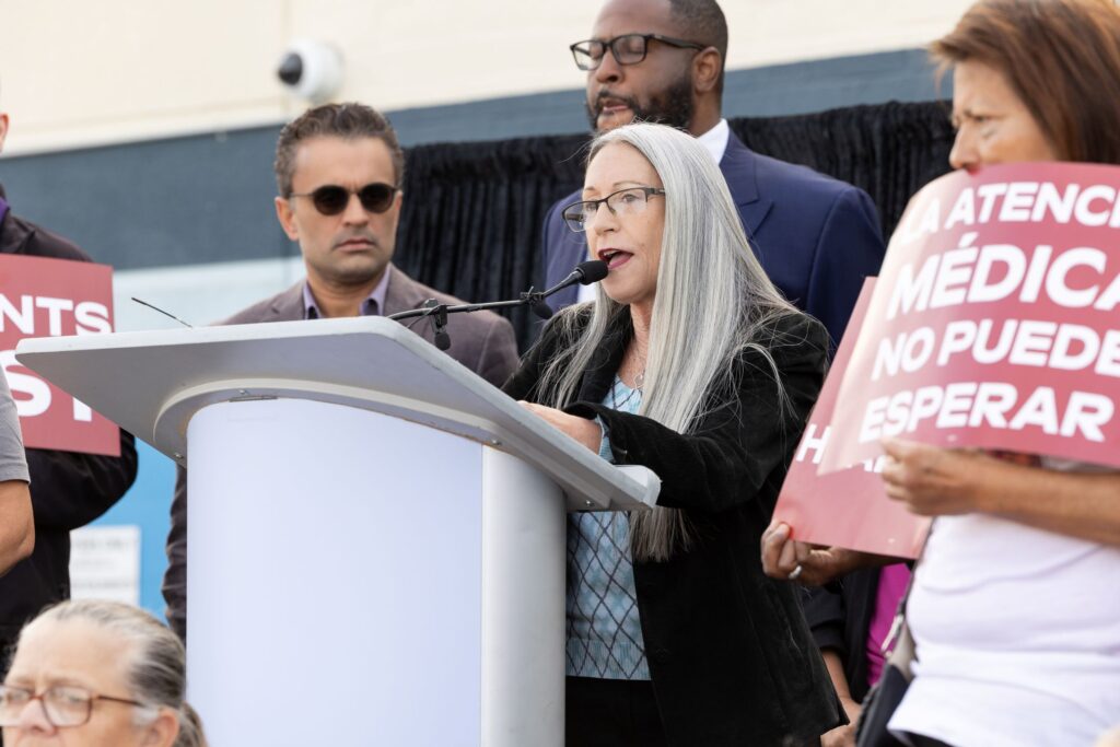 A senior woman speaks at a podium at a rally in support of health care coverage.