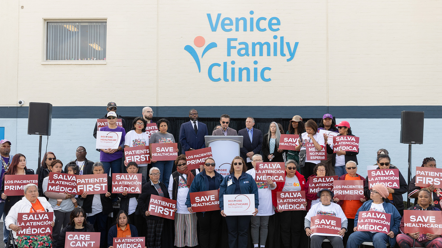 Un Grupo Heterogéneo De Personas De Pie Frente A La Clínica Familiar De Venecia, Con Carteles En Los Que Se Lee SAVE HEALTHCARE Y PATIENTS FIRST En Inglés Y Español, Escuchan A Un Orador En Un Podio.