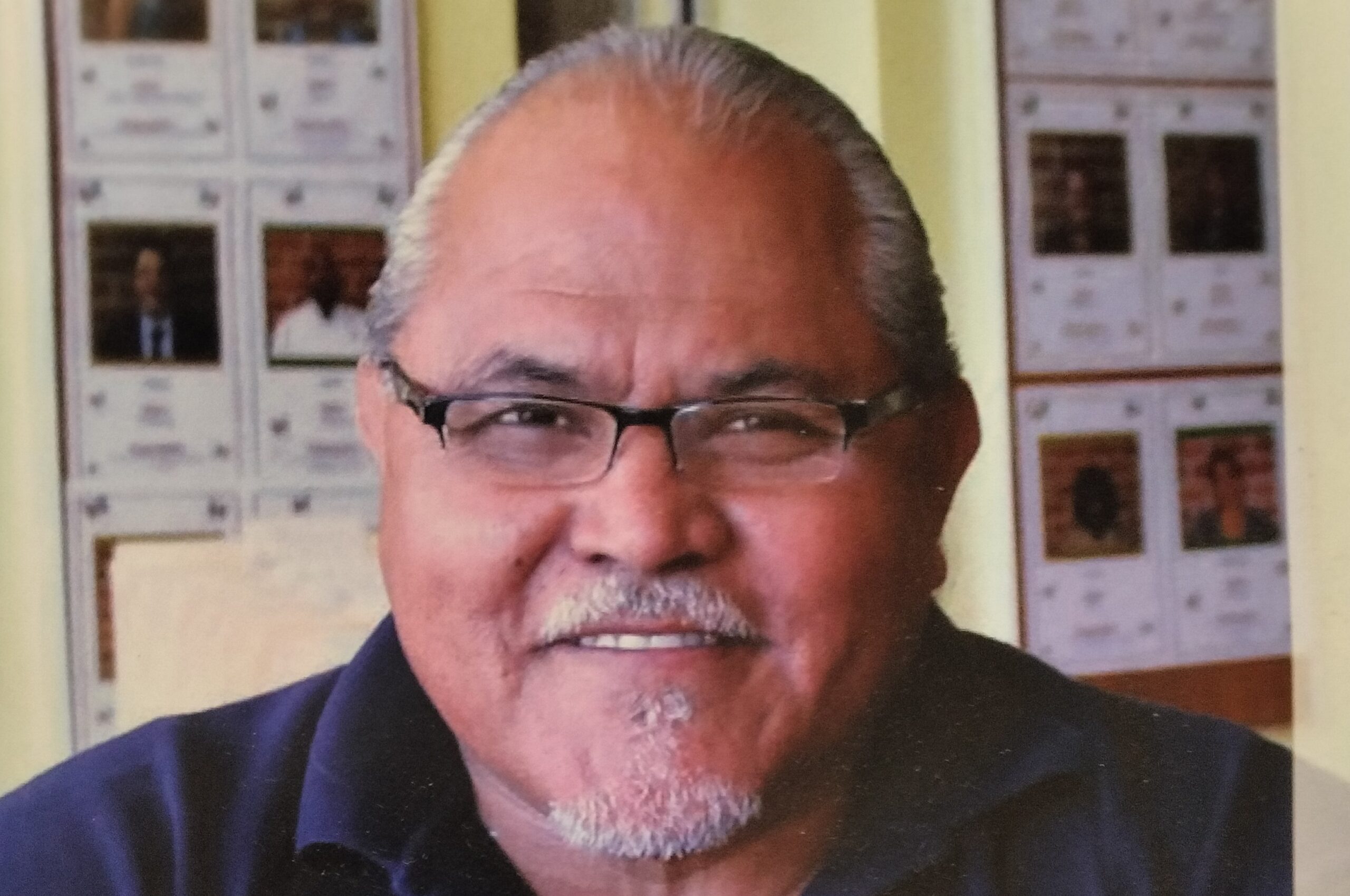 Ernie Rodriguez, a Smiling Older Man With Short Gray Hair, Glasses, And A Goatee, Wearing A Dark Polo Shirt, Sits Indoors With Bulletin Boards Covered In Photos And Documents In The Background.