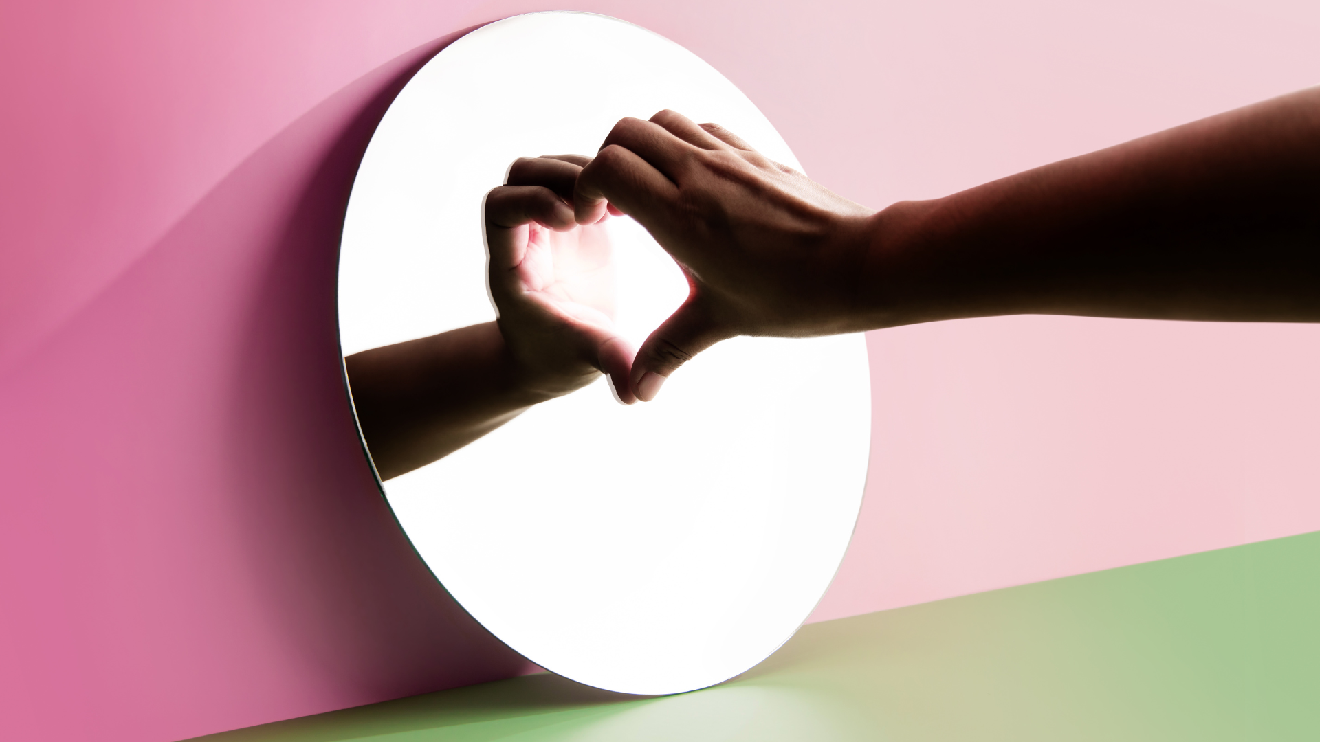 A Person Forms A Heart Shape With Their Hands In Front Of A Round Mirror, With The Reflection Completing The Heart. The Background Features Soft Pink And Green Surfaces. A Person Forms A Heart Shape With Their Hands In Front Of A Round Mirror, With The Reflection Completing The Heart. The Background Features Soft Pink And Green Surfaces.