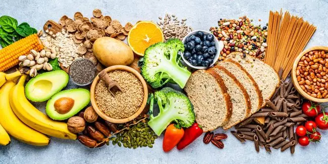 A Colorful Assortment Of Healthy Foods Including Fruits, Vegetables, Whole Grain Bread, Pastas, Nuts, Seeds, And Legumes Arranged On A Light Surface.