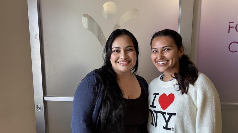 Two Women Stand Side By Side Indoors, Smiling At The Camera. One Wears A Dark Top And The Other Wears A White I ♥ NY Sweatshirt. They Are In Front Of A Frosted Glass Door With A Partial Logo And Text Visible. Two Women Stand Side By Side Indoors, Smiling At The Camera. One Wears A Dark Top And The Other Wears A White I ♥ NY Sweatshirt. They Are In Front Of A Frosted Glass Door With A Partial Logo And Text Visible.