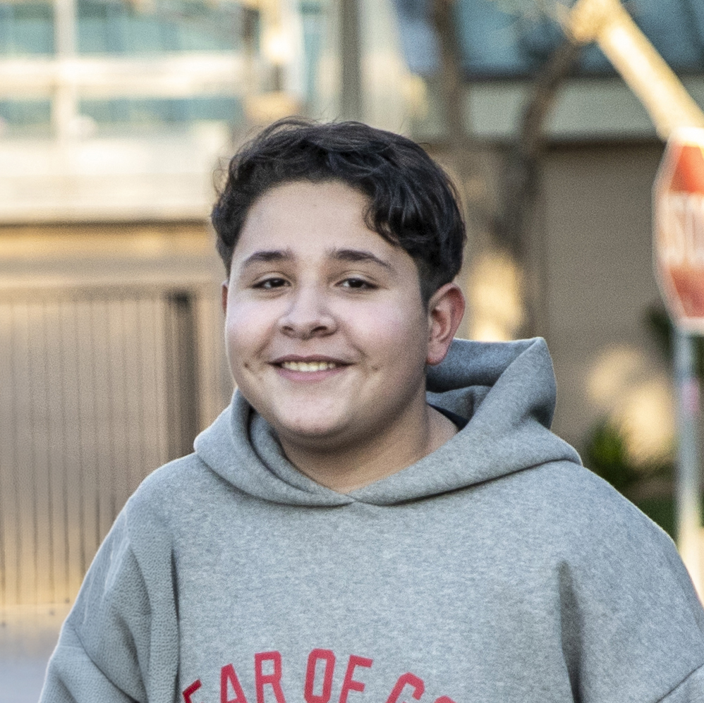 A Smiling Teenage Boy With Short Dark Hair Wears A Gray Hoodie Outdoors. A Blurred Building, Fence, And Stop Sign Are Visible In The Background.