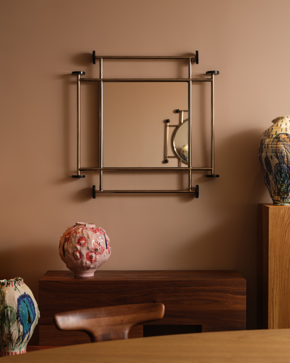 A Square Mirror With A Metal Frame Hangs On A Beige Wall Above A Wooden Cabinet. Colorful, Textured Ceramic Vases And Part Of A Wooden Chair Are Visible In The Foreground.