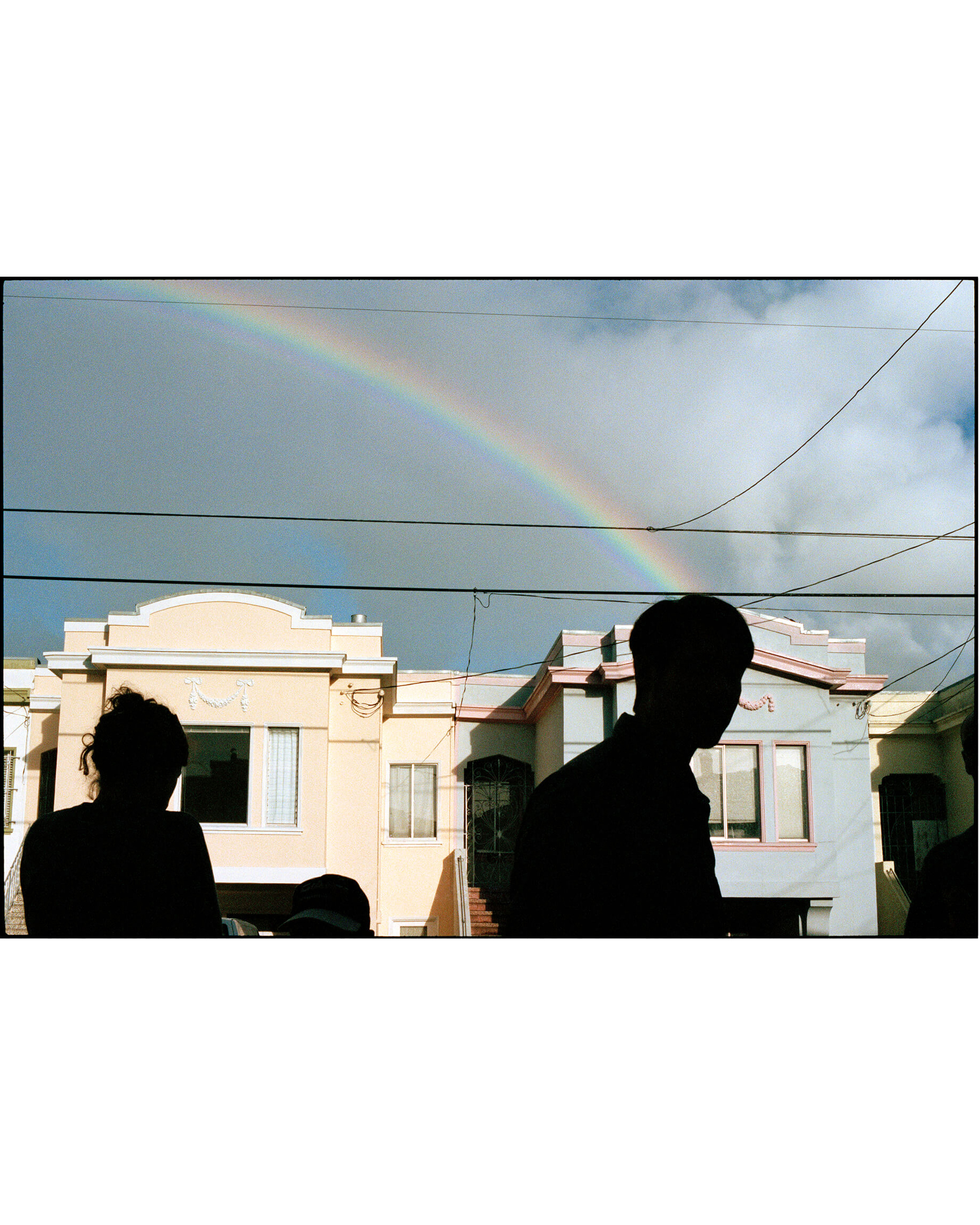 A Rainbow Arcs Across A Cloudy Sky Above Pastel-colored Houses, With The Dark Silhouettes Of Two People In The Foreground.