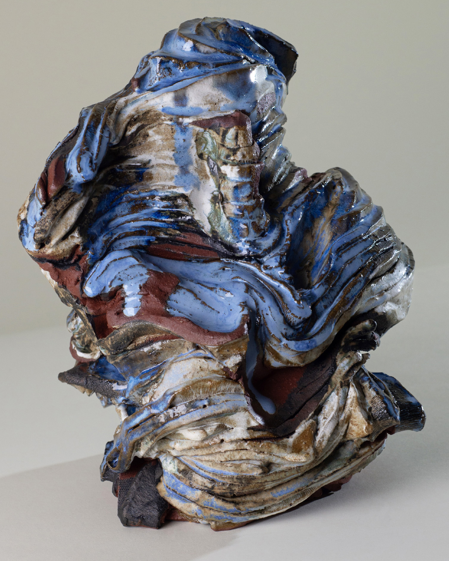 A Sculpted Abstract Ceramic Form Featuring Swirling, Layered Textures In Blue, Brown, White, And Earthy Tones, Resembling Draped Fabric Or Flowing Cloth Against A Plain Background.