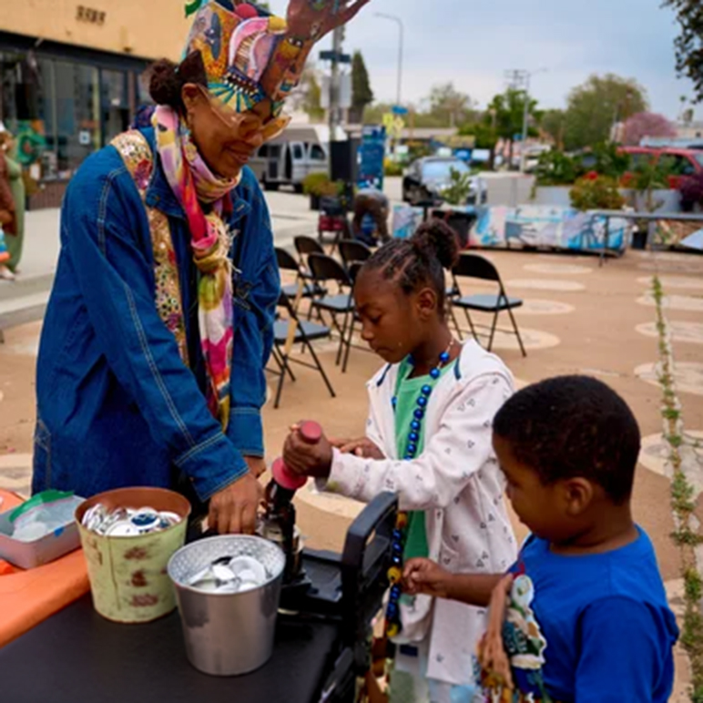 A Woman Wearing A Colorful Hat Helps Two Young Children Use A Machine At An Outdoor Event. There Are Cans In A Bucket On The Table, And Chairs And People In The Background.