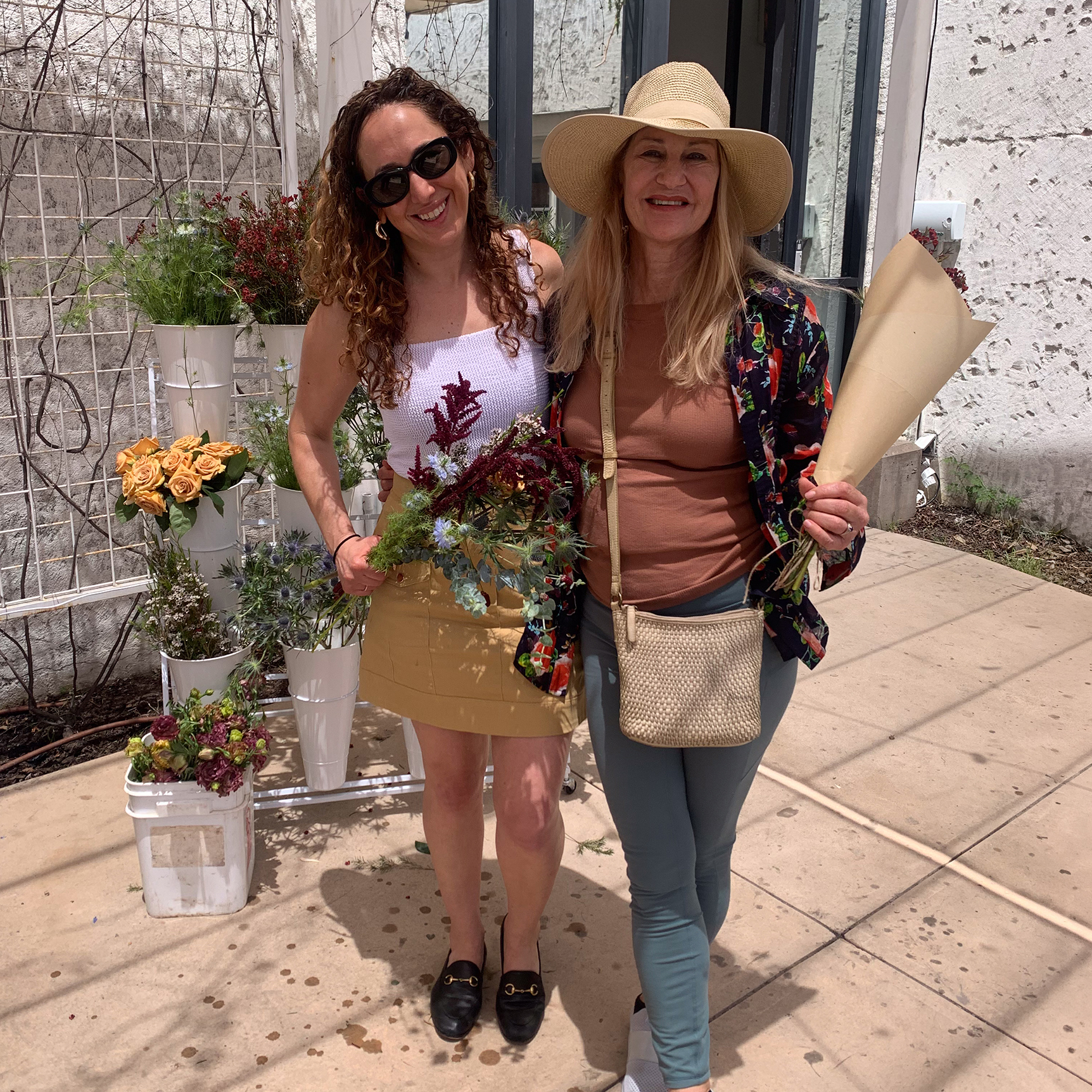 Two Women Smiling Outdoors Near A Flower Stand. One Holds A Bouquet Of Flowers, The Other Holds A Wrapped Bouquet. They Are Casually Dressed And Standing On A Sunny Patio With Potted Flowers Beside Them.