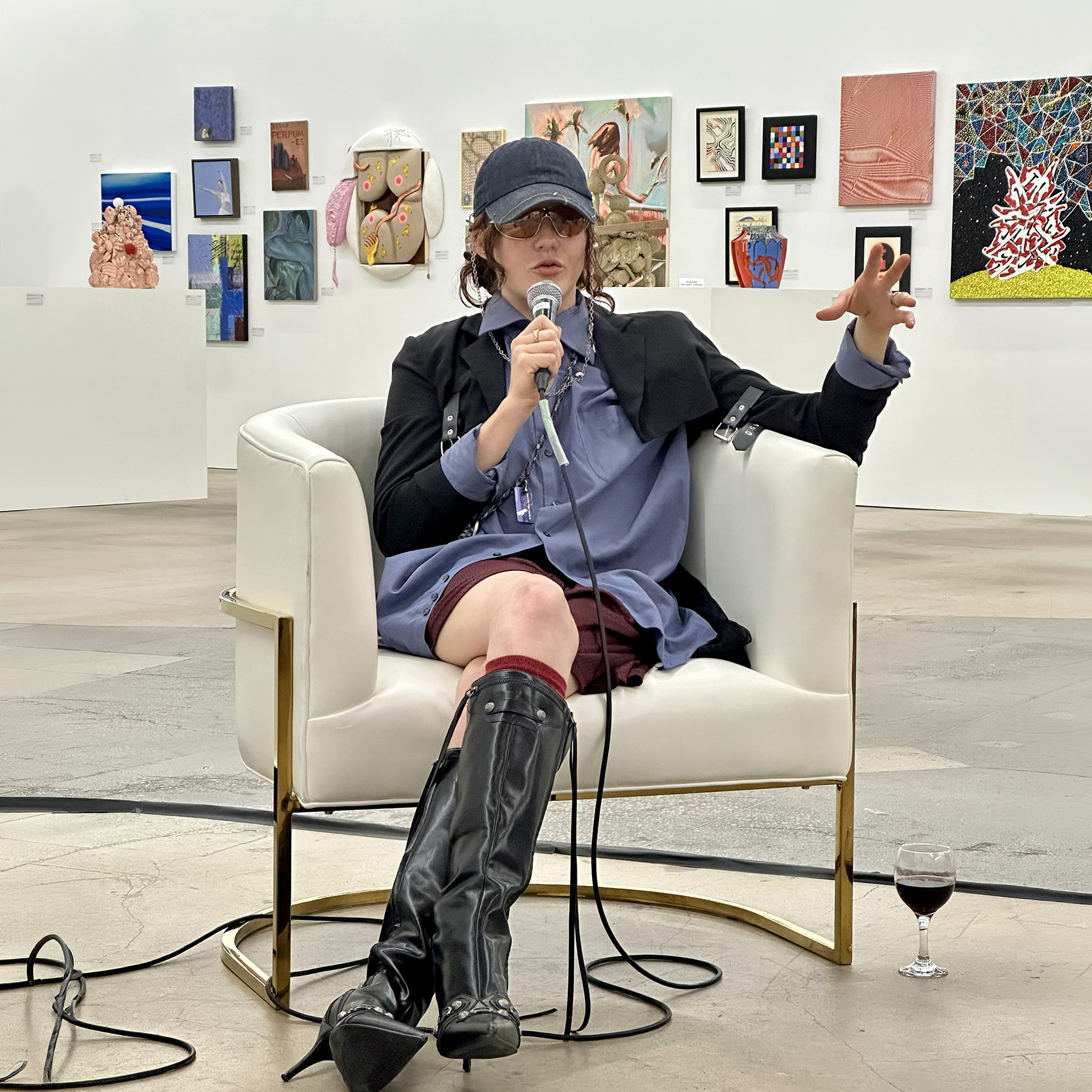 A Person In A Blue Dress, Black Boots, And A Dark Cap Speaks Into A Microphone While Seated In A White Chair At An Art Gallery. Various Colorful Artworks Are Displayed On The Walls Behind Them. A Glass Of Wine Is On The Floor Nearby.