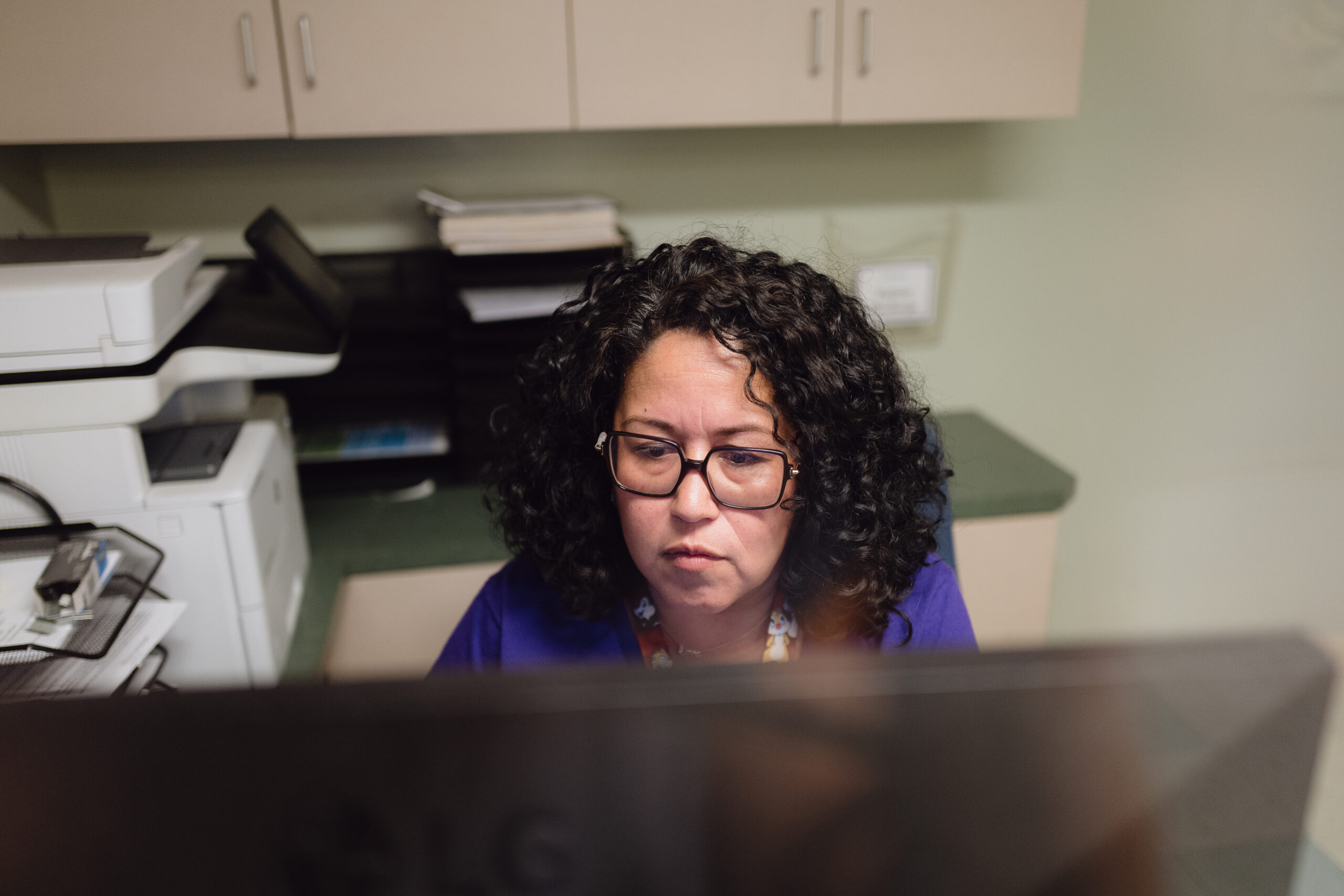 A Woman With Curly Hair And Glasses Works At A Computer In An Office, With Cabinets, Papers, And A Printer In The Background.