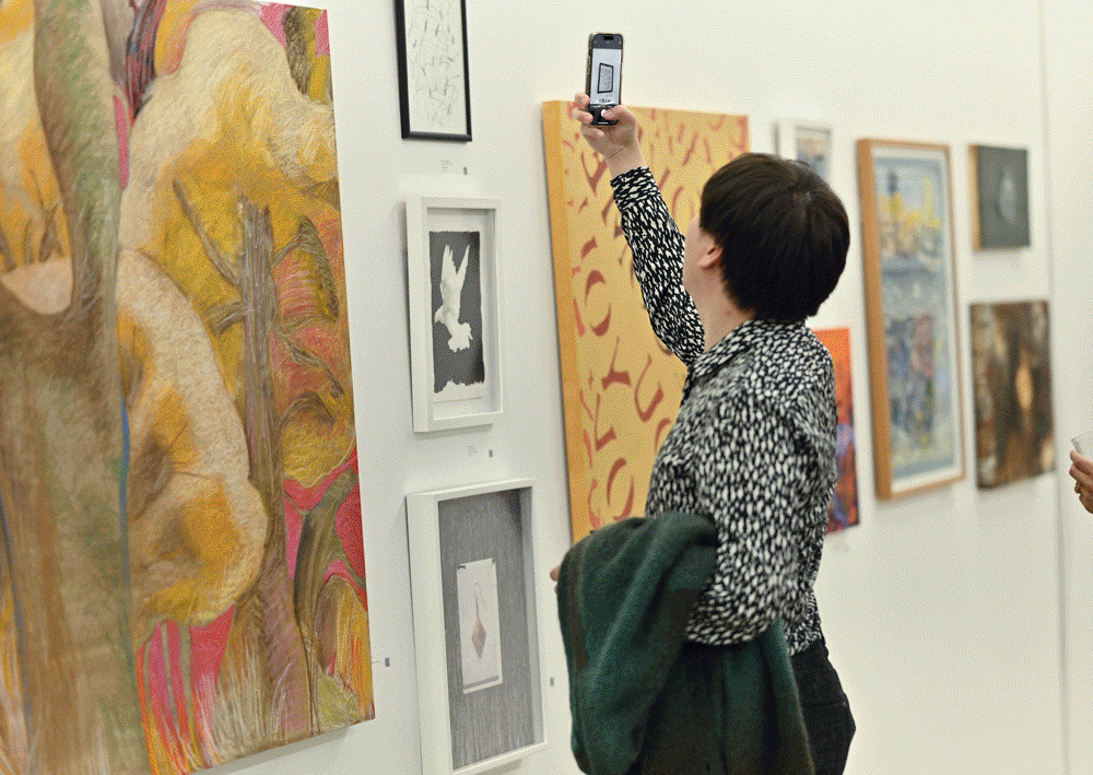 A Person Stands In An Art Gallery, Holding A Coat And Raising Their Phone To Take A Photo Of Artwork On A White Wall Filled With Colorful Paintings And Framed Illustrations.