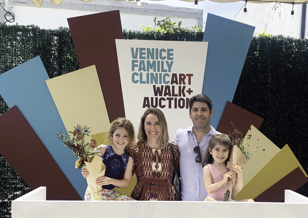 A Smiling Family Of Four Stands In Front Of A Colorful Geometric Backdrop With A Sign Reading “Venice Family Clinic Art Walk + Auction.” Two Children Hold Bouquets Of Flowers, And The Adults Stand Behind Them.