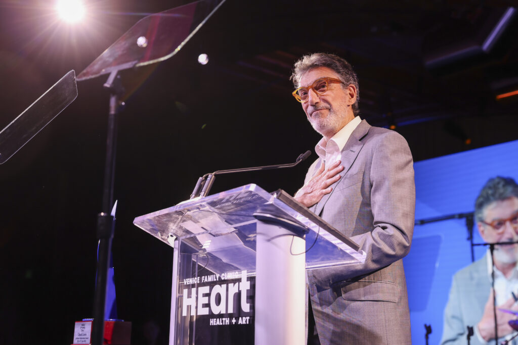 A Man In A Gray Suit Stands At A Clear Podium With The Word “Heart” Displayed On It, Speaking Onstage With One Hand On His Chest. He Is Lit By Stage Lights And Appears Thoughtful, With His Image Projected On A Screen Behind Him.