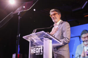 A Man In A Gray Suit Stands At A Clear Podium With The Word “Heart” Displayed On It, Speaking Onstage With One Hand On His Chest. He Is Lit By Stage Lights And Appears Thoughtful, With His Image Projected On A Screen Behind Him.