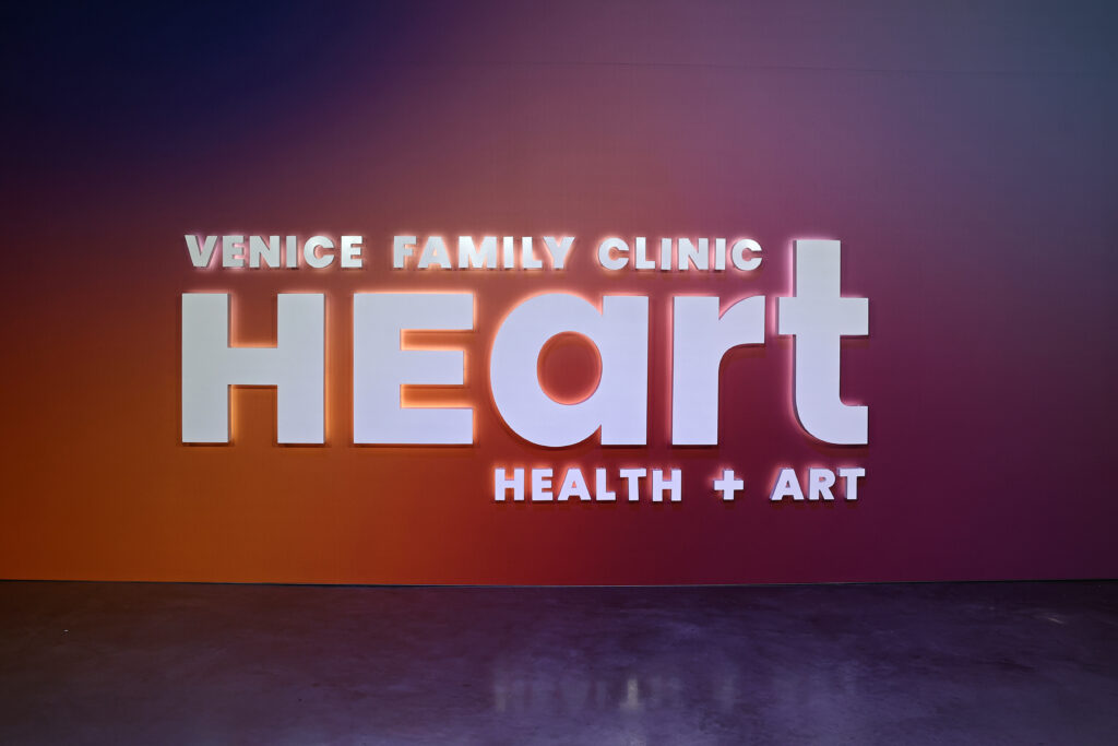 A Wall Displays Large White Text Reading Venice Family Clinic HEART Health + Art Against A Gradient Background That Transitions From Purple To Orange.