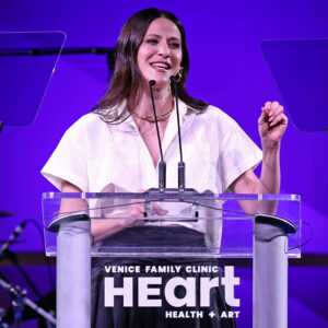 A Woman In A White Shirt Speaks At A Clear Podium With Venice Family Clinic HeART Health + Art Written On The Front. The Background Is Purple, And She Is Smiling.
