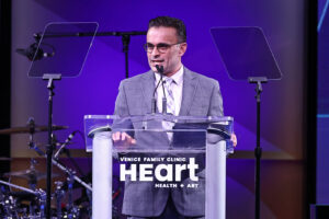 A Man In A Gray Suit And Glasses Speaks At A Clear Podium With The Words Venice Family Clinic Heart Health + Art On The Front. Two Transparent Teleprompters Are Positioned On Either Side Of Him.