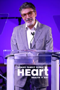 An Older Man With Gray Hair And A Beard, Wearing Glasses And A Gray Suit, Speaks At A Clear Podium Labeled Venice Family Clinic Heart Health + Art Against A Purple-lit Background.